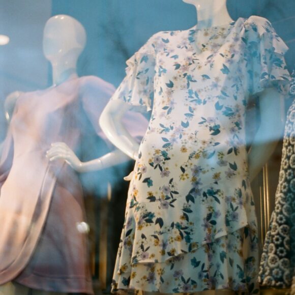Mannequins in a shop window display floral and pastel dresses. The reflection on the glass adds a dreamy, elegant tone to the scene.