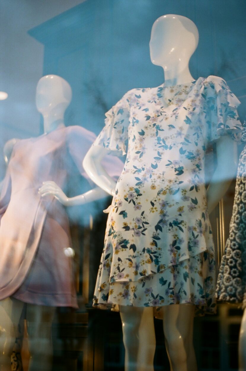 Mannequins in a shop window display floral and pastel dresses. The reflection on the glass adds a dreamy, elegant tone to the scene.