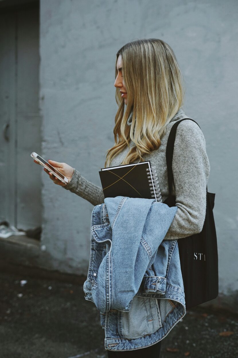 The Fashion-Forward Pack That Actually Works A woman with long hair stands outdoors, holding a smartphone, notebook, and denim jacket. She looks focused, wearing a gray sweater and black bag. Urban setting.