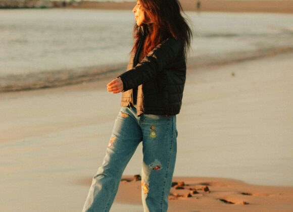 ๐ Pacific Coast Highway Road Trip: A Stylish Journey in Your Pacific Coast Jeans A woman with long hair walks barefoot on a sandy beach at sunset, leaving footprints in the wet sand. She wears a black jacket and denim jeans.