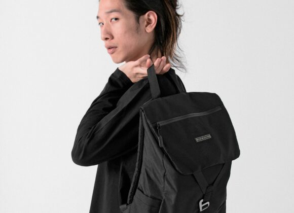 🌟 Longchamp Backpack Review: Style Meets Functionality Person standing against a white background, looking over their shoulder while holding a black backpack. They have long hair tied back, wearing a dark outfit.