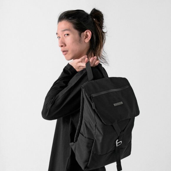 Person standing against a white background, looking over their shoulder while holding a black backpack. They have long hair tied back, wearing a dark outfit.