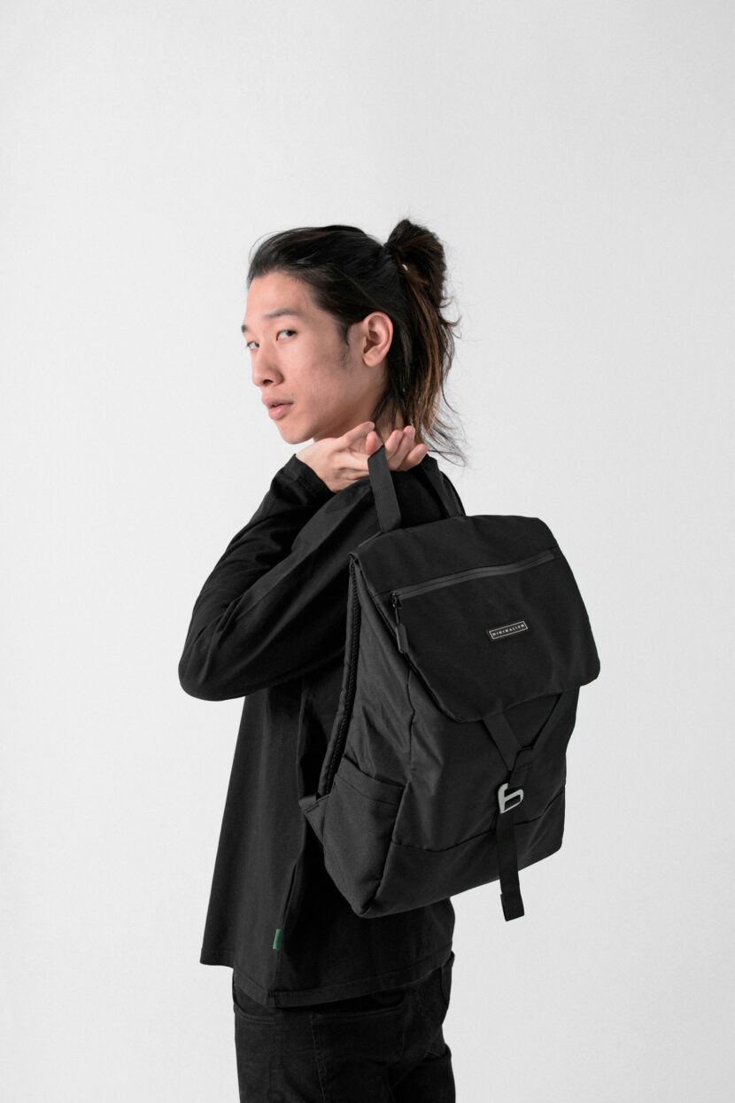 Person standing against a white background, looking over their shoulder while holding a black backpack. They have long hair tied back, wearing a dark outfit.