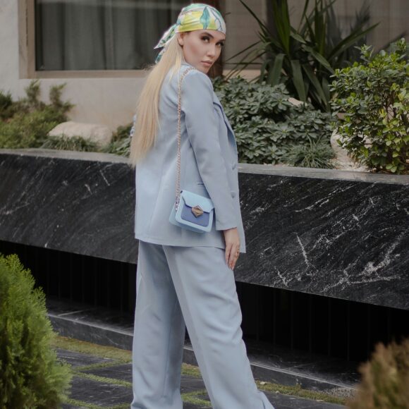 A person in a light blue suit and white sneakers stands outside. They wear a colorful headscarf and carry a matching light blue purse, exuding confidence.