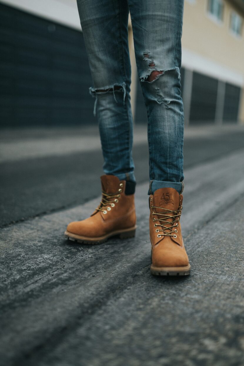 👢 Best Mid-Calf Boots for Women: Style Guide for Pairing Jeans with Mid-Calf Boots A person wearing distressed jeans and tan work boots stands on a dark, textured road. The background features blurred garages, creating an urban feel.