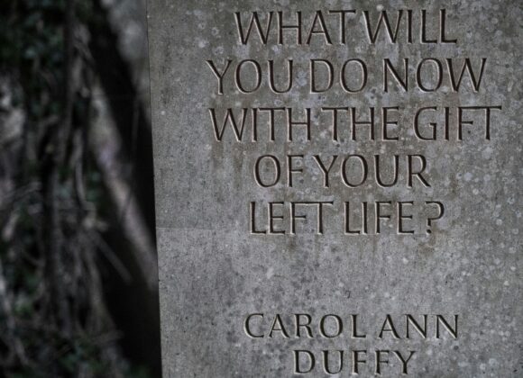Stone slab with carved words: "What will you do now with the gift of your left life? Carol Ann Duffy." Background shows blurred foliage, creating a contemplative mood.
