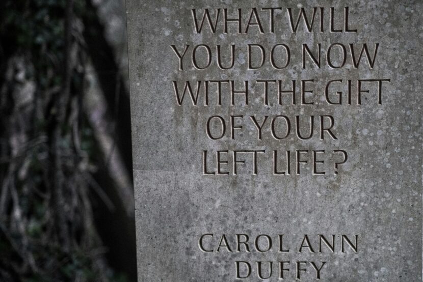 Stone slab with carved words: "What will you do now with the gift of your left life? Carol Ann Duffy." Background shows blurred foliage, creating a contemplative mood.
