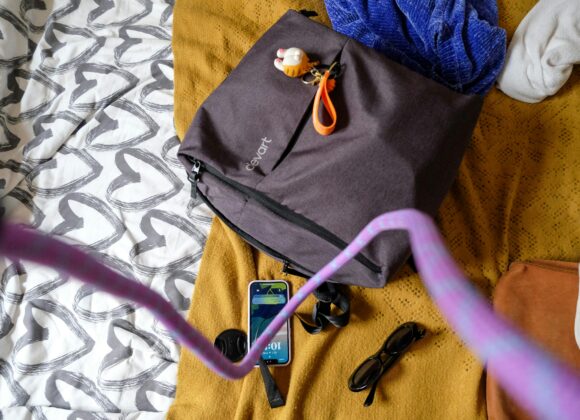 The Ultimate Bali Fashion & Packing Guide A brown bag with a keychain and a purple strap on a multicolored bedspread. Nearby are a smartphone, sunglasses, and a lens cap, conveying a casual and ready-to-go vibe.