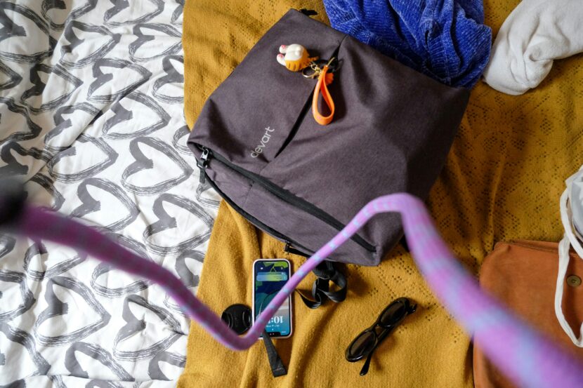 The Ultimate Bali Fashion & Packing Guide A brown bag with a keychain and a purple strap on a multicolored bedspread. Nearby are a smartphone, sunglasses, and a lens cap, conveying a casual and ready-to-go vibe.