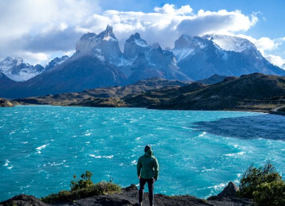10-Step Packing Guide for South America: Smart, Stylish & Climate-Ready A person in a green jacket stands by a vibrant turquoise lake, gazing at rugged snow-capped mountains under a partly cloudy sky, evoking adventure.