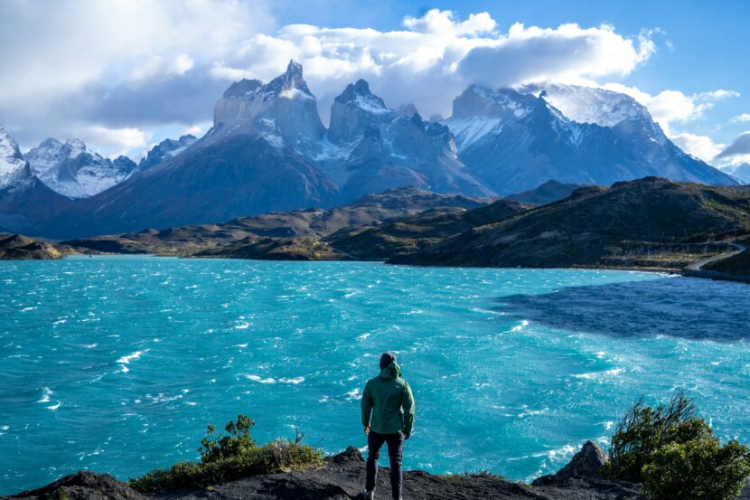 10-Step Packing Guide for South America: Smart, Stylish & Climate-Ready A person in a green jacket stands by a vibrant turquoise lake, gazing at rugged snow-capped mountains under a partly cloudy sky, evoking adventure.