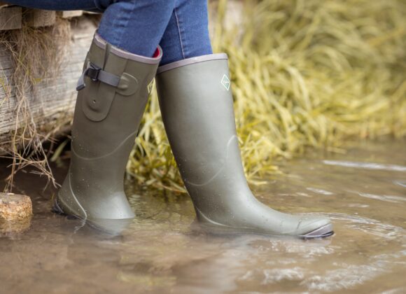 🥾 Best Waterproof Boots for Women and Men Person in green rubber boots relaxes by a shallow stream, with one boot in the water. They are seated on a wooden structure, surrounded by grass.