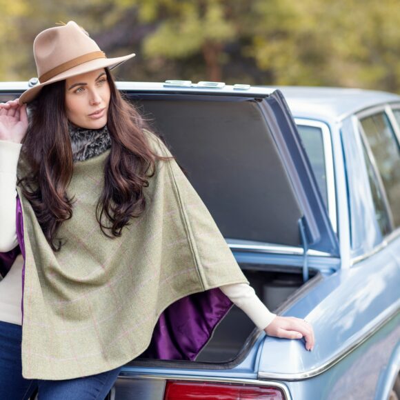 A woman in a beige hat and green poncho leans against an open car trunk. She gazes thoughtfully to the side. The background features blurred greenery.