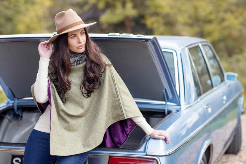 Reversible Clothing for Travel A woman in a beige hat and green poncho leans against an open car trunk. She gazes thoughtfully to the side. The background features blurred greenery.