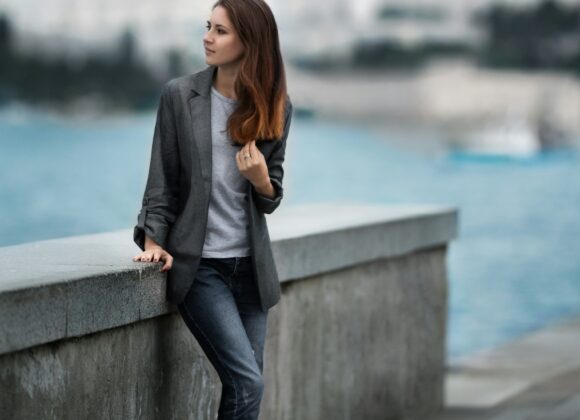 Best Travel Jeans for Women: Your Ultimate Guide to Comfort, Style, and Functionality Young woman in a gray blazer and jeans leans against a stone wall by the sea. The background is a soft-focus cityscape, conveying a calm, reflective mood.