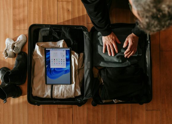 ✈️ The Ultimate List of Toiletries for Travel: Pack Smart, Stay Fresh A person packs clothes into an open suitcase on a wooden floor, with a tablet displaying a screen inside. Nearby are boots and white sneakers. The scene feels organized and ready for travel.