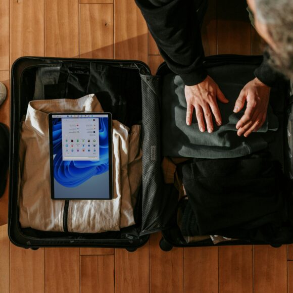 A person packs clothes into an open suitcase on a wooden floor, with a tablet displaying a screen inside. Nearby are boots and white sneakers. The scene feels organized and ready for travel.