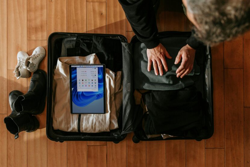 A person packs clothes into an open suitcase on a wooden floor, with a tablet displaying a screen inside. Nearby are boots and white sneakers. The scene feels organized and ready for travel.