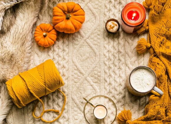 Cozy autumn scene with a cream sweater, small pumpkins, a candle, a hot drink, yellow yarn, and an orange blanket, evoking warmth and comfort.