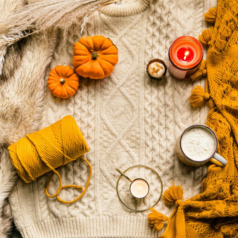 What to Wear in NYC in October, November & Winter Cozy autumn scene with a cream sweater, small pumpkins, a candle, a hot drink, yellow yarn, and an orange blanket, evoking warmth and comfort.