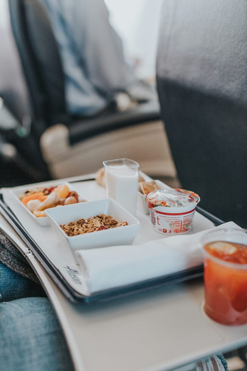 ✈️ Best Snacks for Flights That Keep You Full and Happy Airplane tray with breakfast items, including granola, yogurt, juice, and a glass of milk, set on a passenger's tray table. Cozy travel ambiance.