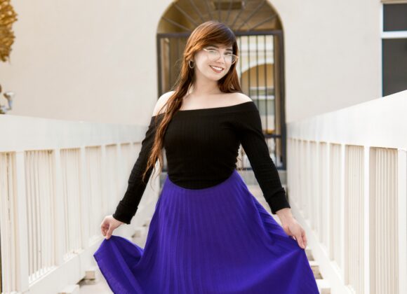 Elevate Your Style with Black and Purple Accents A woman in a black off-the-shoulder top and bright purple skirt stands on a bridge, smiling. Palm leaves frame the scene, evoking a cheerful atmosphere.