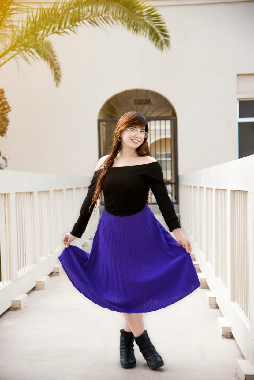 Elevate Your Style with Black and Purple Accents A woman in a black off-the-shoulder top and bright purple skirt stands on a bridge, smiling. Palm leaves frame the scene, evoking a cheerful atmosphere.