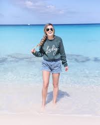 What to Wear in Turks and Caicos A woman joyfully walks on a beach, wearing sunglasses, a green "Lucky" sweater, and denim shorts. Crystal-clear turquoise water and blue sky set a serene scene.