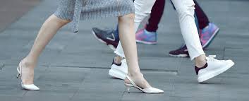 Women's Narrow Shoes: The Ultimate Guide to Fit People walking on a gray sidewalk, featuring close-up of legs. One person wears white heels and a gray skirt, others in casual sneakers and pants. Urban scene.