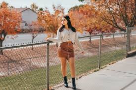 ✨ The Ultimate Guide to Women’s Midi Skirts A woman in a white sweater and brown skirt stands by a chain-link fence. She's surrounded by autumn trees with orange leaves, under a bright sky.