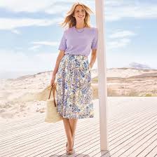The Timeless Charm of Women's Linen Skirts Woman in a lavender shirt and floral skirt stands on a wooden deck, holding a straw bag. Background of sandy dunes and blue sky conveys a relaxed, summery vibe.