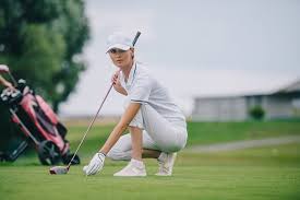 ⛳ Tee Off in Style: The Ultimate Guide to Cute Golf Dresses for Women A woman golfer in a white outfit and cap crouches on a green, aligning her putt. A golf bag is in the background. The scene conveys focus and concentration.