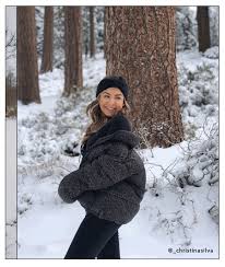 Winter Travel Outfits with American Eagle Outfitters Woman smiling in snowy forest, wearing a dark fluffy coat and black beanie. Snow blankets the ground and trees, creating a peaceful winter scene.