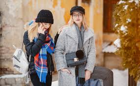 Winter Accessories for Women: Style Meets Warmth Two women in winter attire stand outside. One wears a black hat and scarf, looking at her phone. The other wears a gray coat and black cap, holding a handbag. Snow is on the ground.