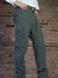 đš The Curious Case of Convertible Travel Pants Person wearing dark gray cargo pants with multiple pockets, standing against a rustic brick wall. The tone is casual and utilitarian.