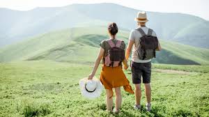 Packing for a Trip: When to Start and How Long It Really Takes A couple with backpacks stands on a vibrant green hill, facing distant mountains. The woman holds a white hat, creating a serene and adventurous tone.