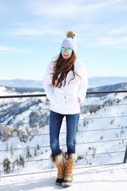 What to Wear in the Snow A woman in a white jacket, jeans, and fur boots stands on a snowy mountain overlook. She wears a blue beanie and sunglasses, projecting a cheerful, wintry vibe.