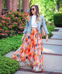 š§„ What to Wear with a Maxi Skirt A woman in a flowy, colorful maxi skirt and denim jacket walks on a garden path. She wears sunglasses and carries a shoulder bag, surrounded by greenery.