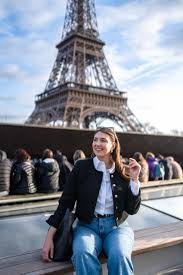 What to Wear to Paris A woman smiling and sitting on a ledge with the Eiffel Tower in the background under a blue sky. She wears a black jacket and jeans, exuding happiness.