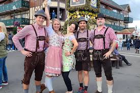 What to Wear to Oktoberfest in Munich A group of five people in traditional Bavarian attire, smiling at a festival. Half-timbered buildings and a maypole in the background. Festive and joyful atmosphere.