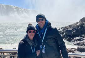 What to Wear to Niagara Falls Couple dressed warmly, smiling in front of a misty waterfall under a clear blue sky. They're standing near rocks with a joyful and adventurous vibe.