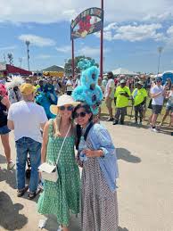 What to Wear to New Orleans Two women in summer dresses and hats smile at an outdoor festival. Behind them, a crowd enjoys the sunny day. A sign and blue sky are visible above.