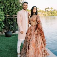 🕌 What to Wear to an Indian Wedding A couple in elegant attire, the man in a pale pink sherwani and the woman in a flowing, patterned gown, stand by a serene lakeside with lush greenery.