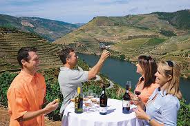 What to Wear on a Rhine River Cruise A group of four people enjoy wine around a table with bottles and glasses, set against a scenic backdrop of lush hills and a winding river. They seem relaxed and happy.