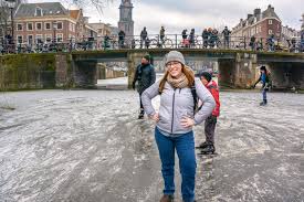 What to Wear in the Netherlands A person in a gray jacket and beanie smiles while standing on a snowy path. A bridge and several people are in the background, creating a lively, wintry scene.