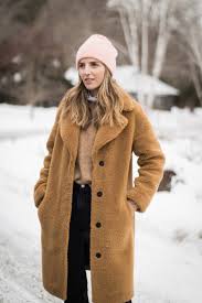 What to Wear in Winter A woman in a brown coat and pink beanie stands in a snowy landscape. Bare trees and a snow-covered house create a peaceful, wintry backdrop.