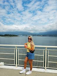 What to Wear in Vancouver A smiling person in sunglasses and casual summer clothes leans on a waterfront railing, holding a cup. The sky is partly cloudy, evoking a relaxed atmosphere.