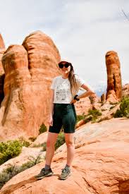 What to Wear in Utah Summer A woman wearing a white t-shirt, shorts, and cap stands on red rocky terrain with large rock formations in the background, conveying adventure and exploration.