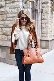What to Wear in the Midwest A woman in sunglasses and a white sweater, draped with a brown jacket, walks on a city street, smiling and carrying a large tan handbag.