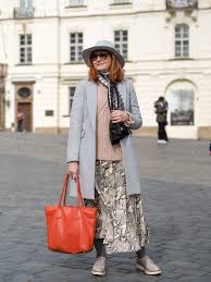 What to Wear in the Czech Republic A woman in chic streetwear stands in a square, wearing a gray coat, patterned skirt, and hat, holding a bright orange bag, exuding a fashionable and confident vibe.